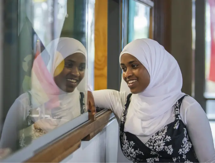 A dark-skinned person leaning on a reception counter with their face reflected in the glass. 