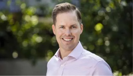 A headshot of BSL Executive Director Travis McLeod wearing a light pink dress shirt and standing outside with trees behind him.