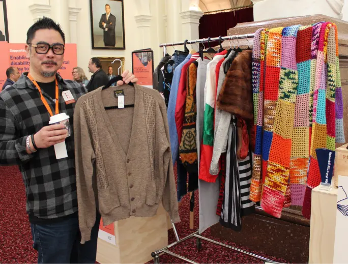 BSL staff member Justin Tan is pictured at the BSL expo. They are standing holding a brown cardigan in their left hand and a take away coffee in their right hand.