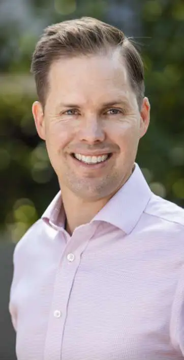 A headshot of BSL Executive Director Travis McLeod wearing a light pink dress shirt and standing outside with trees behind him.