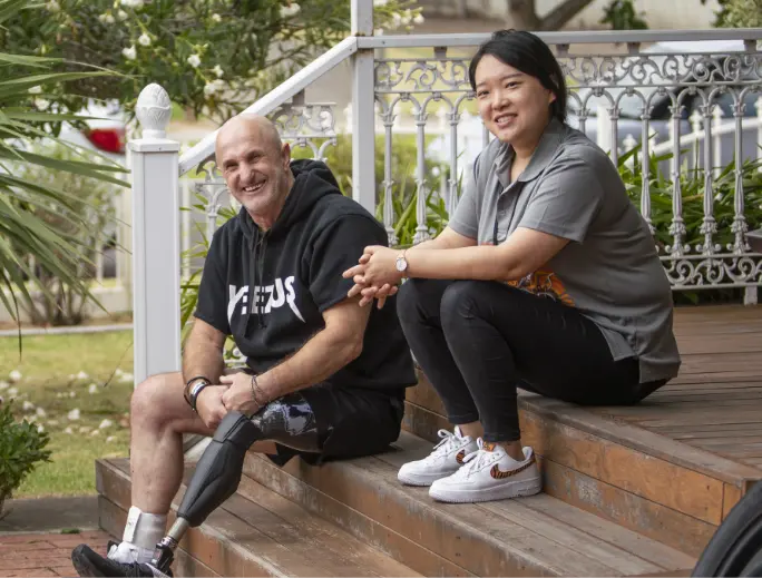 BSL NDIS participant Tim and Local Area Coordinator Yi are sitting on a step outside what looks like a house, enjoying a break after a workout.